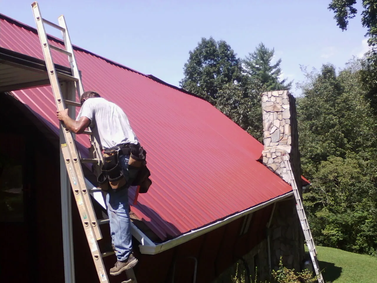 Roofer on a ladder beside a red metal roof and stone chimney — Soffit & Fascia Repair by Roofing Brothers in Tallmadge